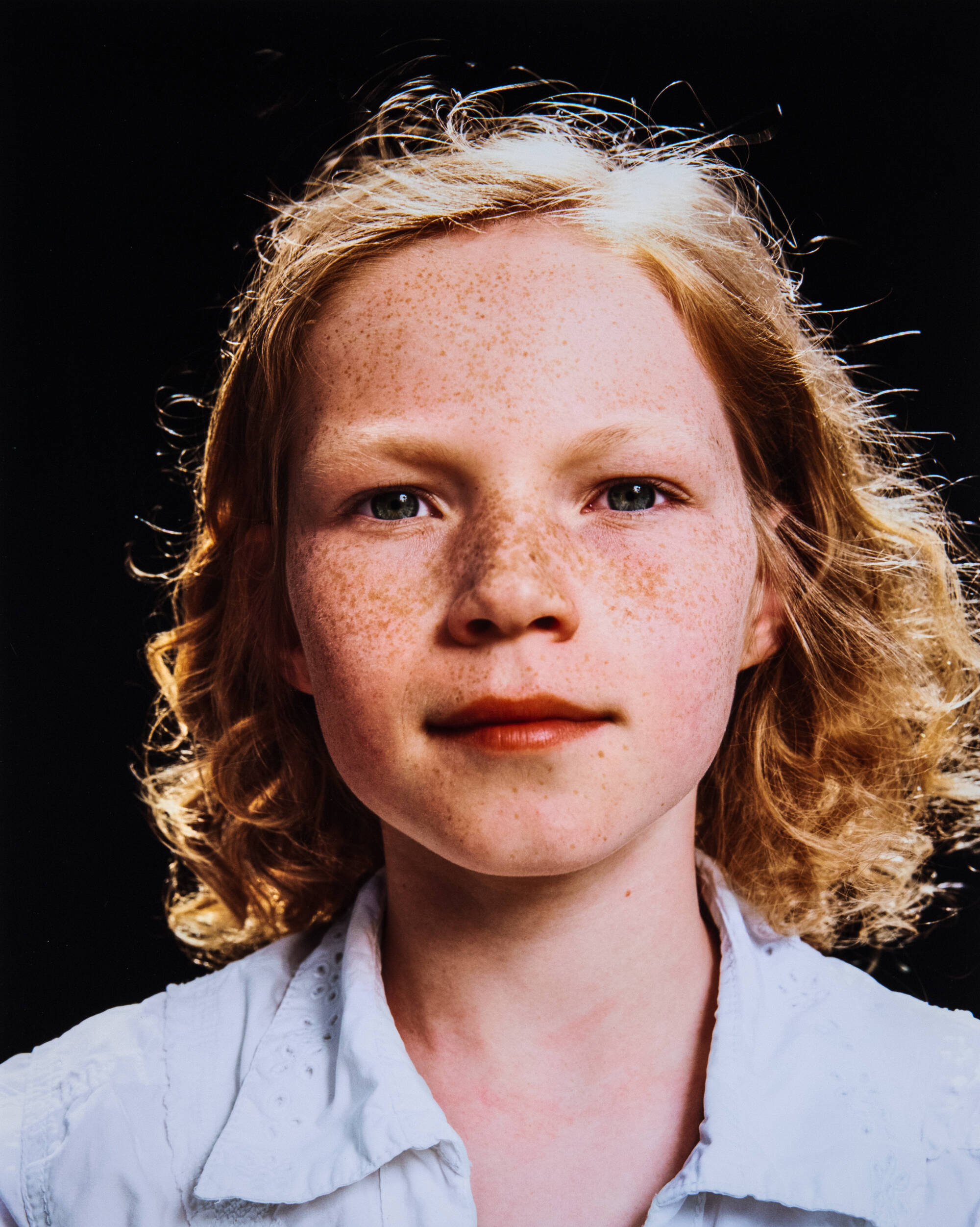Color photograph of a child's portrait. They have curly hair, freckles, and wear a light blue shirt.